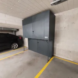 Szara szafa garażowa na metalowym stelażu zamykana na klucz, umieszczona w garażu obok zaparkowanego czarnego samochodu.