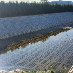 Wybetonowany rów melioracyjny z ażurowymi płytami, częściowo wypełniony wodą, z widocznym odbiciem ażurowej konstrukcji w tafli wody, otoczony zielenią.