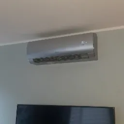 Szary klimatyzator marki Gree zamontowany na ścianie w pokoju, widoczny wyświetlacz temperatury 22 stopnie, pod nim czarny telewizor.