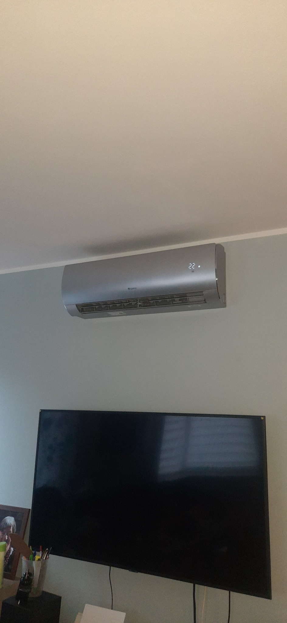 Szary klimatyzator marki Gree zamontowany na ścianie w pokoju, widoczny wyświetlacz temperatury 22 stopnie, pod nim czarny telewizor.