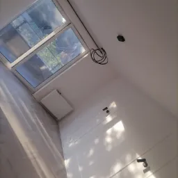 Wykończone poddasze z oknem dachowym, białymi ścianami z dekoracyjnymi listwami, grzejnikiem pod oknem i parkietem ułożonym w jodełkę, częściowo oświetlone promieniami słonecznymi.