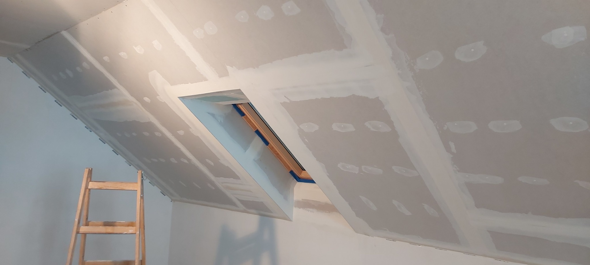 Poddasze z oknem dachowym w trakcie prac wykończeniowych, widoczne płyty gipsowo-kartonowe ze spoinami wypełnionymi masą szpachlową, drewniana drabina oparta o ścianę.