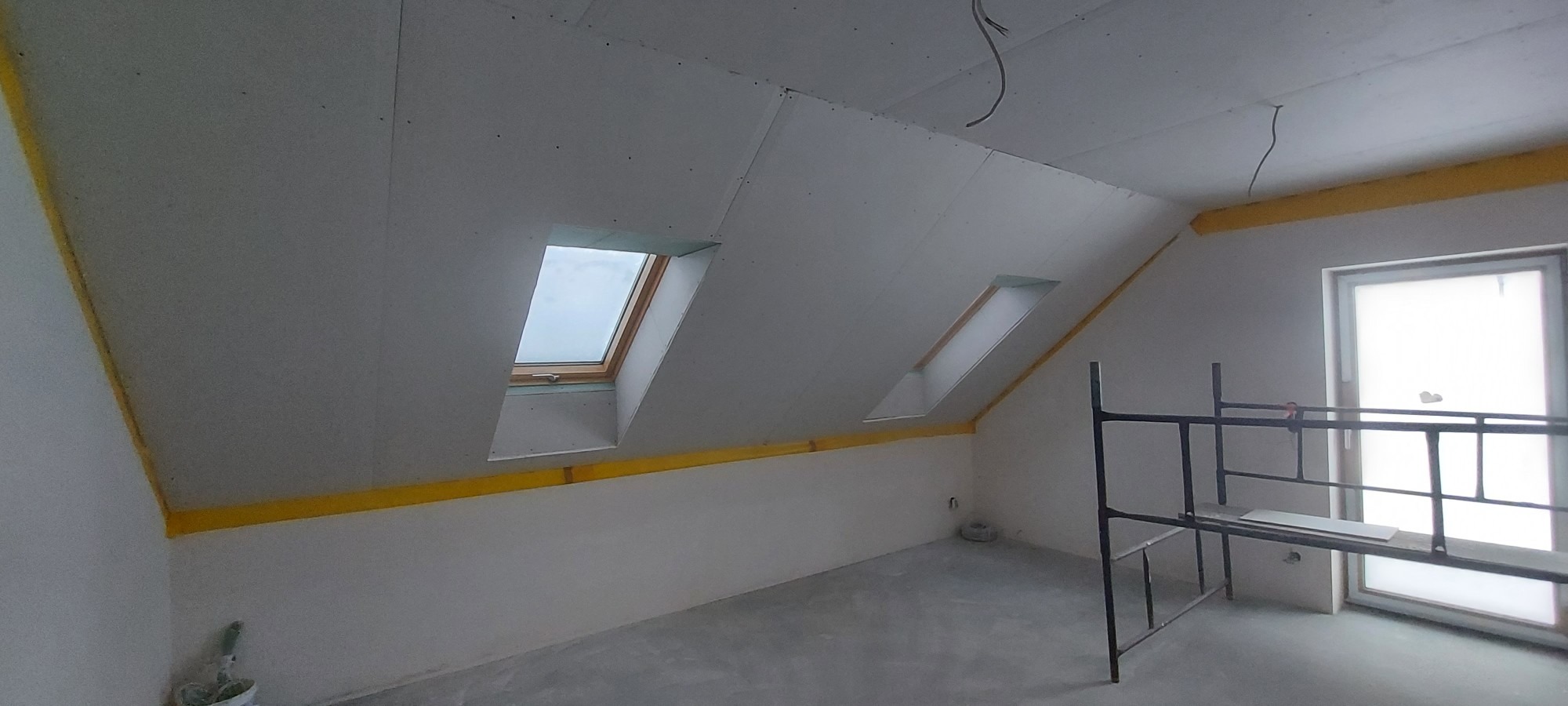 Poddasze z zamontowanymi płytami gipsowo-kartonowymi na skosach i suficie, widoczne okna dachowe i rusztowanie malarskie, przygotowane do dalszych prac wykończeniowych.