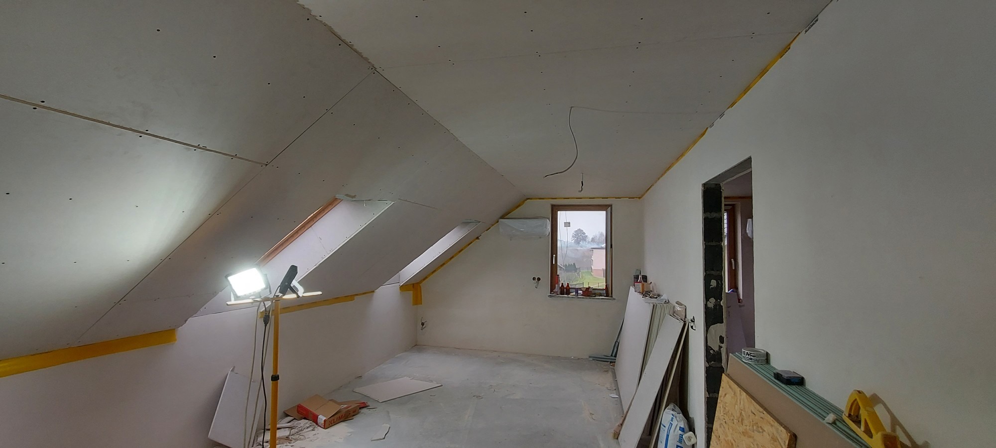 Poddasze w trakcie wykańczania: zamontowane płyty gipsowo-kartonowe na skośnym suficie i ścianach, widoczne okna dachowe i tymczasowe oświetlenie halogenowe, przygotowane materiały budowlane...
