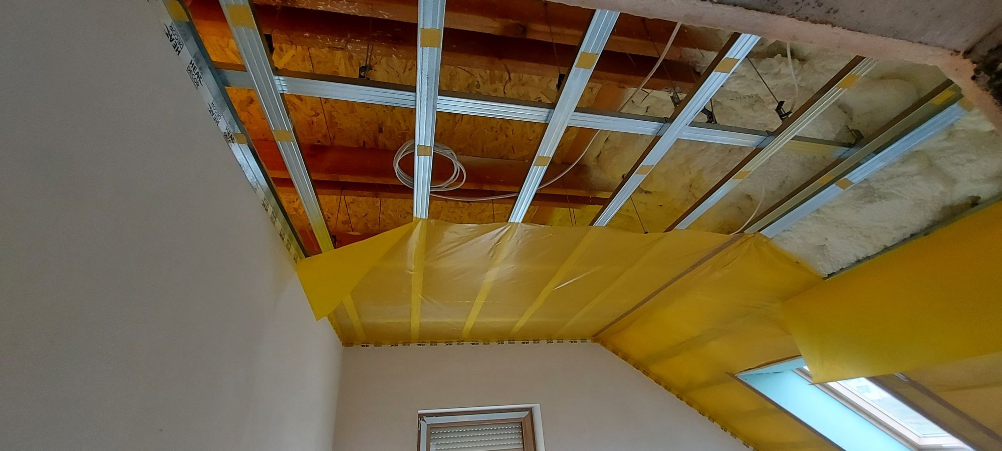 Sufit poddasza w trakcie prac wykończeniowych: widoczne metalowe profile, izolacja z wełny mineralnej, folia ochronna w kolorze żółtym i fragment ściany. Okno dachowe w tle.