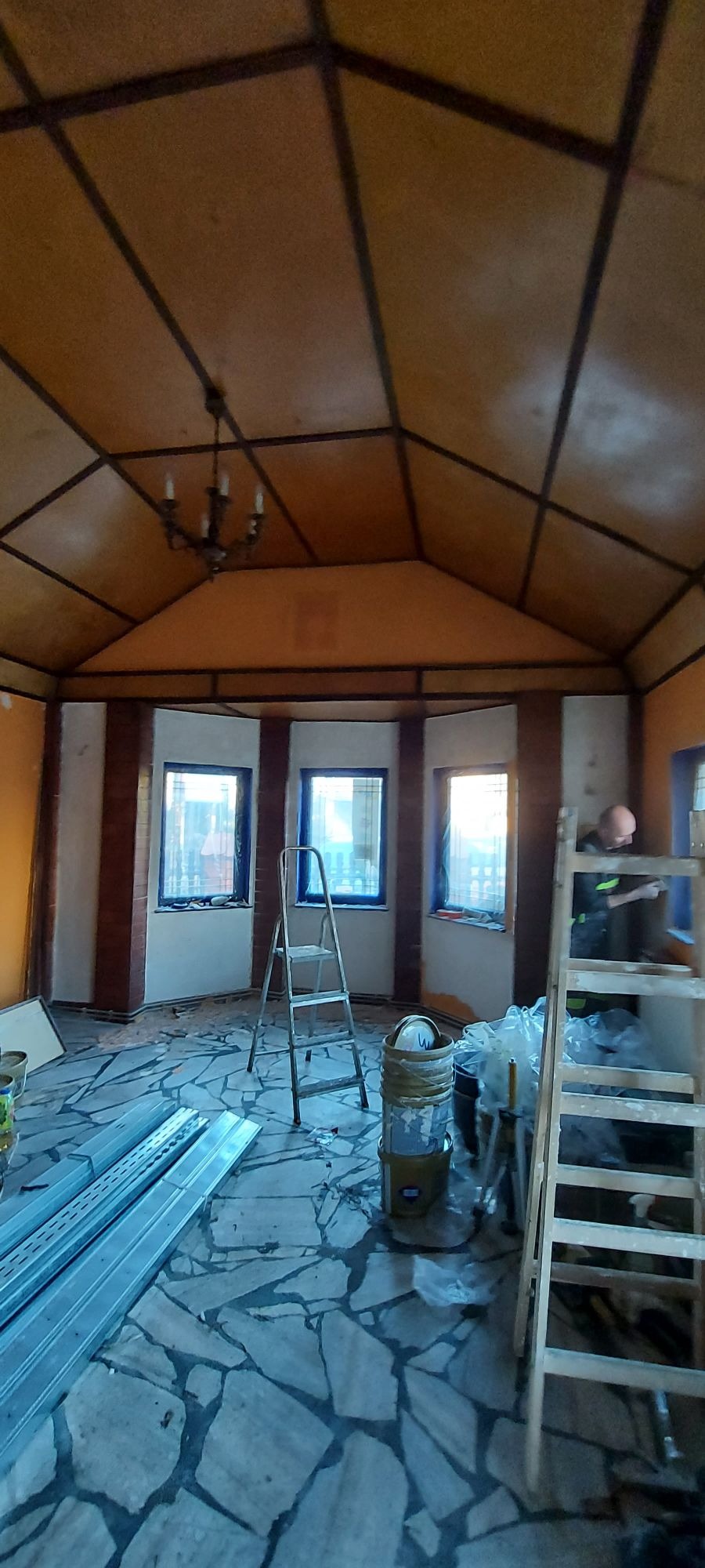 Malowanie wnętrza pokoju o nietypowej konstrukcji sufitu: widoczne okna zabezpieczone folią, drabiny, puszki z farbą i pracownik malujący ramę okienną.