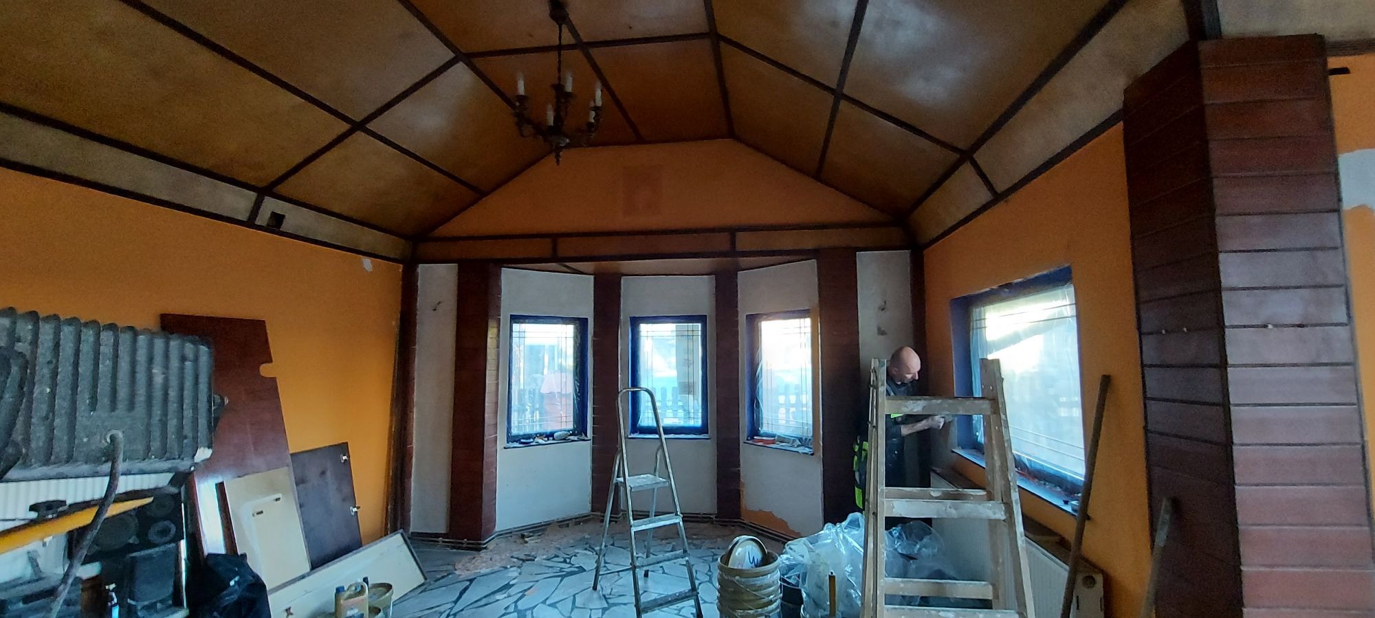 Wnętrze remontowanego pokoju z pomarańczowymi ścianami, drewnianym stropem i mozaikową podłogą. Pracownik na drabinie przy oknie, zabezpieczone okna folią, widoczne narzędzia i materiały budowlane.