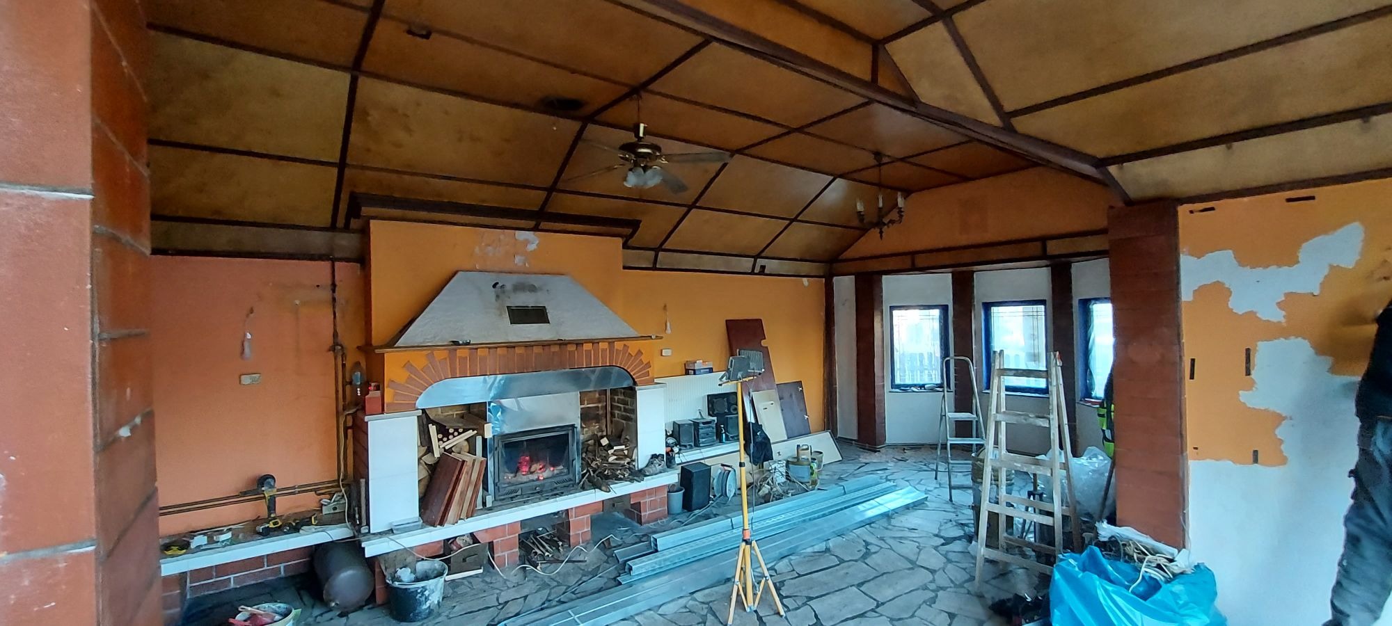 Wnętrze pokoju z kominkiem w trakcie remontu; widoczne ślady po wcześniejszym malowaniu ścian, metalowe profile na podłodze, drabina i sprzęt oświetleniowy.