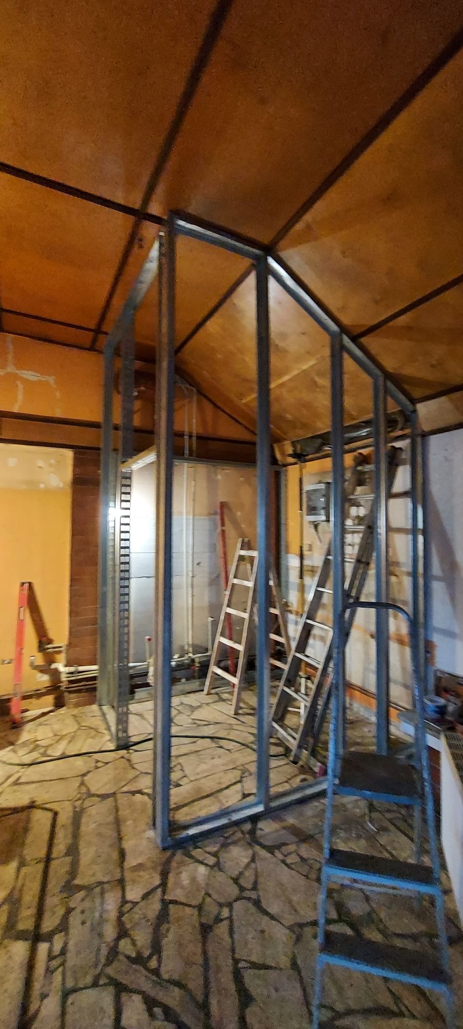 Stelaż z profili stalowych w trakcie budowy ścianki działowej, widoczne drabiny i fragmenty instalacji w surowym wnętrzu z drewnianą boazerią na suficie.