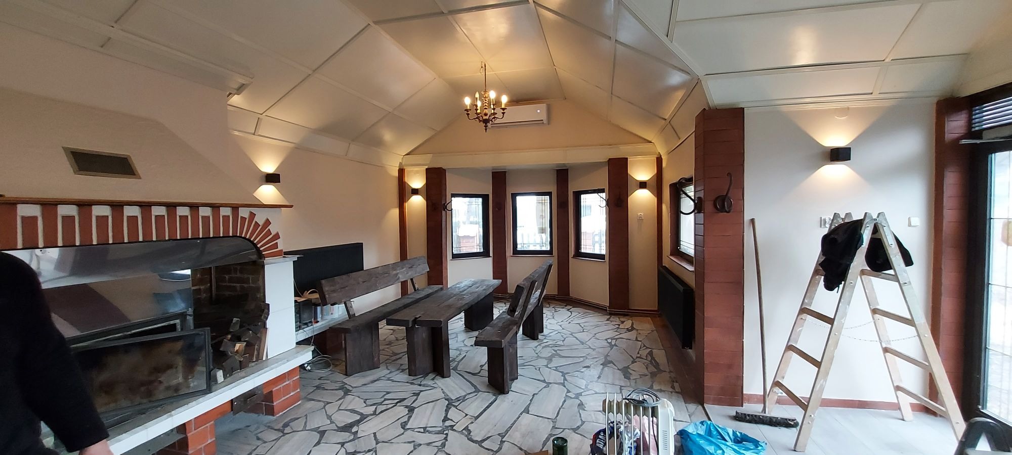 Przestronne wnętrze z marmurową posadzką, drewnianymi meblami, kominkiem z cegły i białymi ścianami, oświetlone przez nowoczesne kinkiety i żyrandol. Na ścianie widoczny klimatyzator, a w rogu...