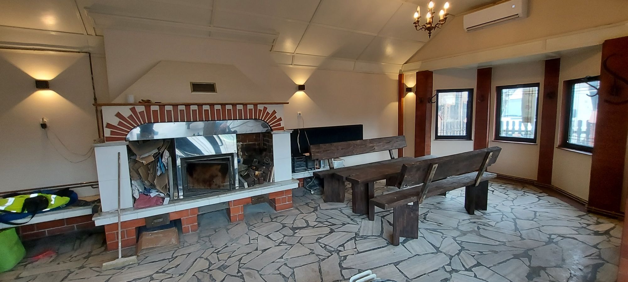 Wnętrze z kominkiem obłożonym cegłą i stalą nierdzewną, kamienna posadzka, drewniane ławy i stoły, oświetlenie punktowe i żyrandol, widoczny klimatyzator.