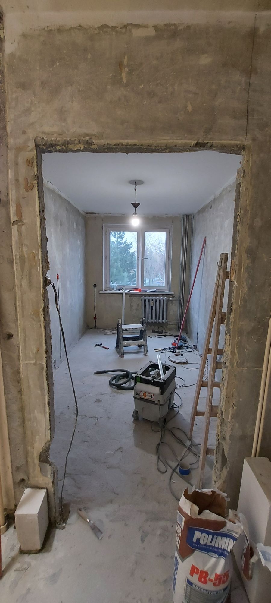 Pokój w trakcie remontu, ściany przygotowane do dalszych prac, widoczny worek z zaprawą, drabina i odkurzacz przemysłowy.