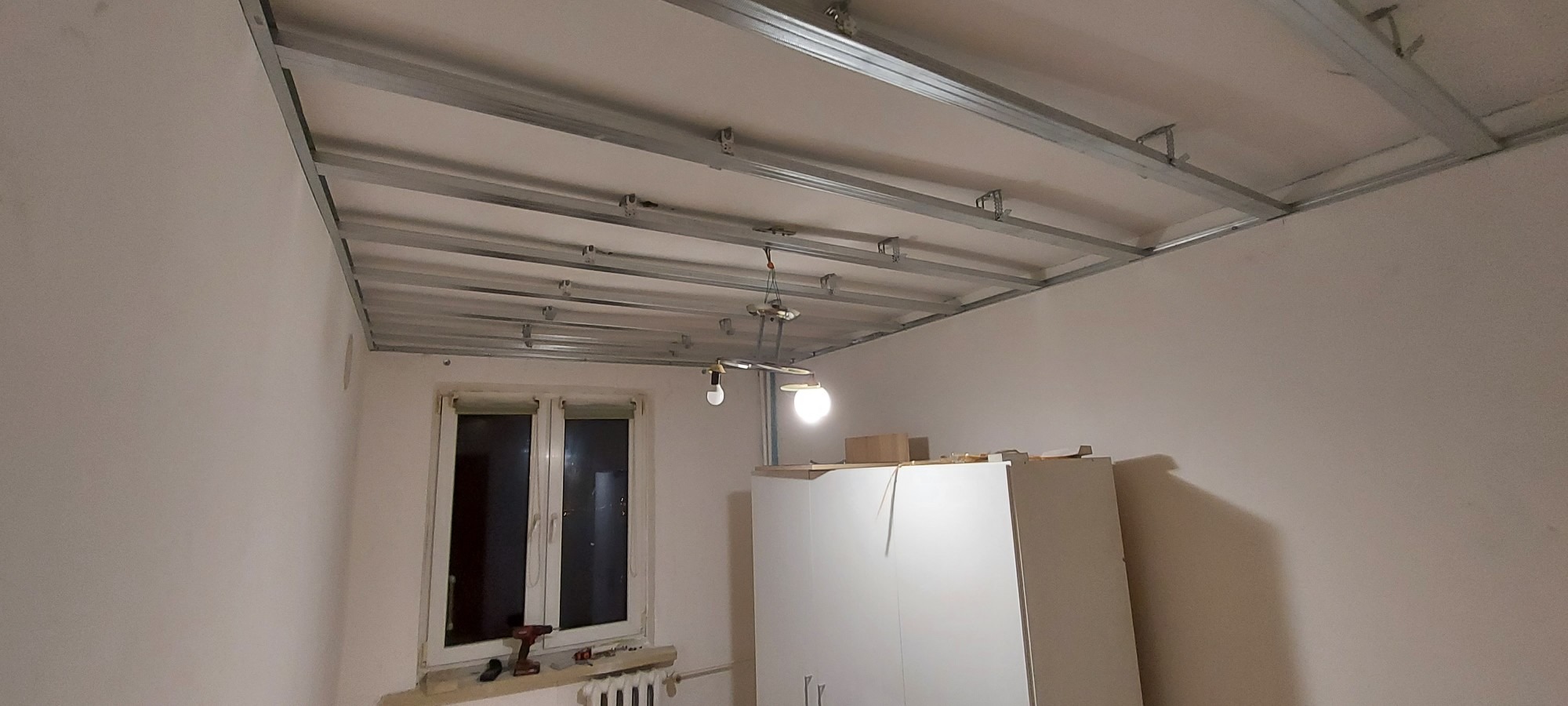 Montaż metalowej konstrukcji sufitu podwieszanego w pokoju z oknem, widoczna instalacja elektryczna i narzędzia na parapecie.