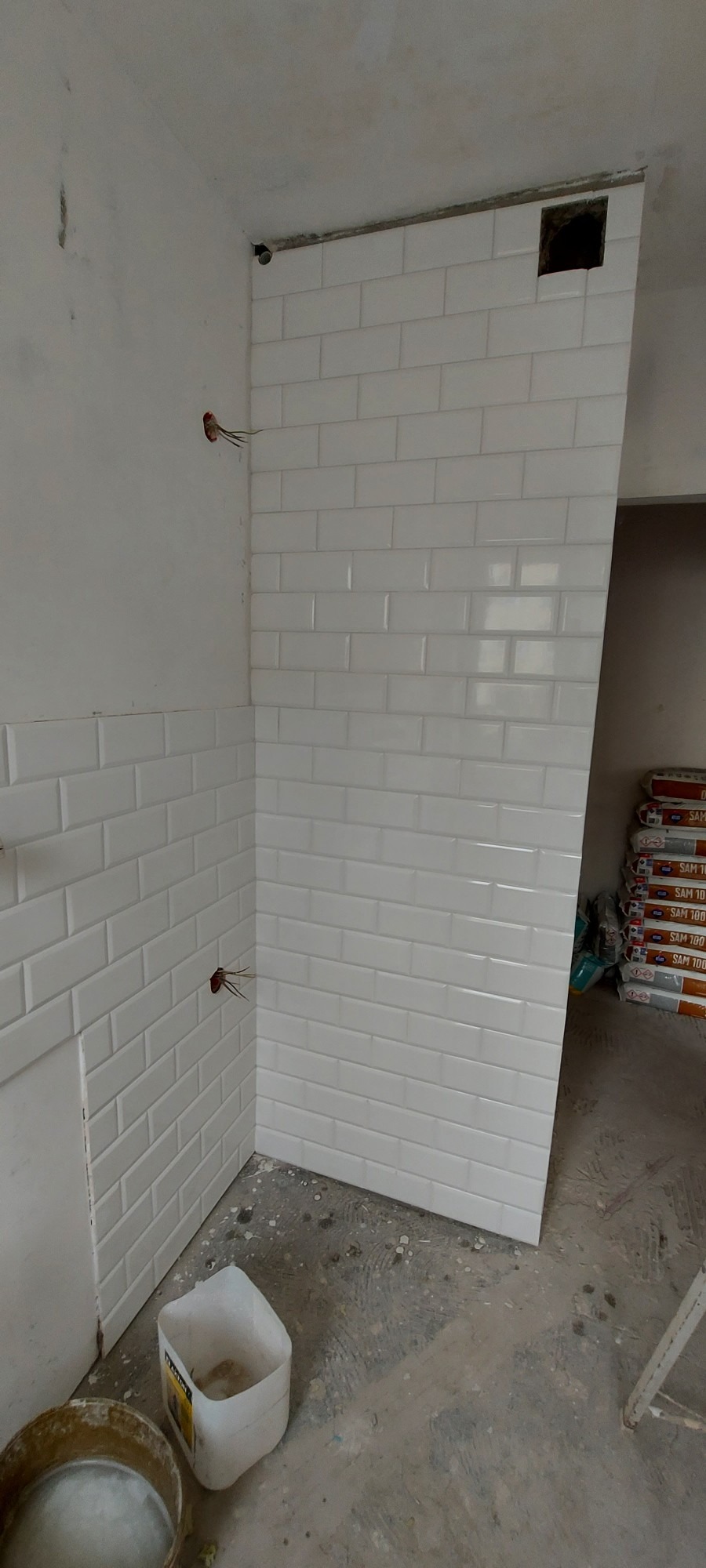 Świeżo ułożone, białe, prostokątne płytki ścienne typu metro w rogu pomieszczenia, widoczne wystające kable elektryczne i worki z zaprawą w tle.