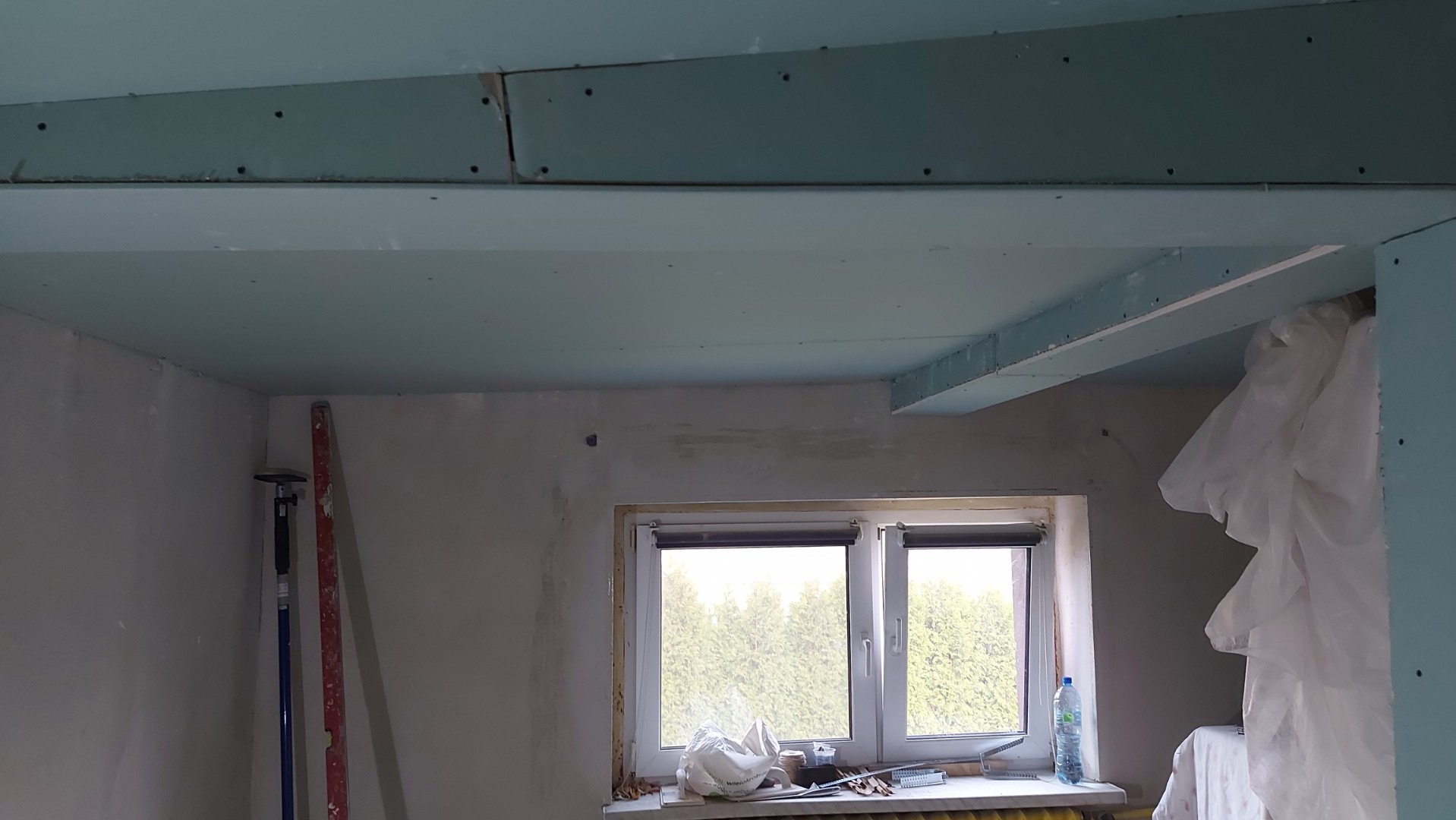 Wnętrze remontowanego pokoju z częściowo zamontowanymi płytami gipsowo-kartonowymi na suficie i ścianach, widoczne narzędzia i butelka wody na parapecie okna.