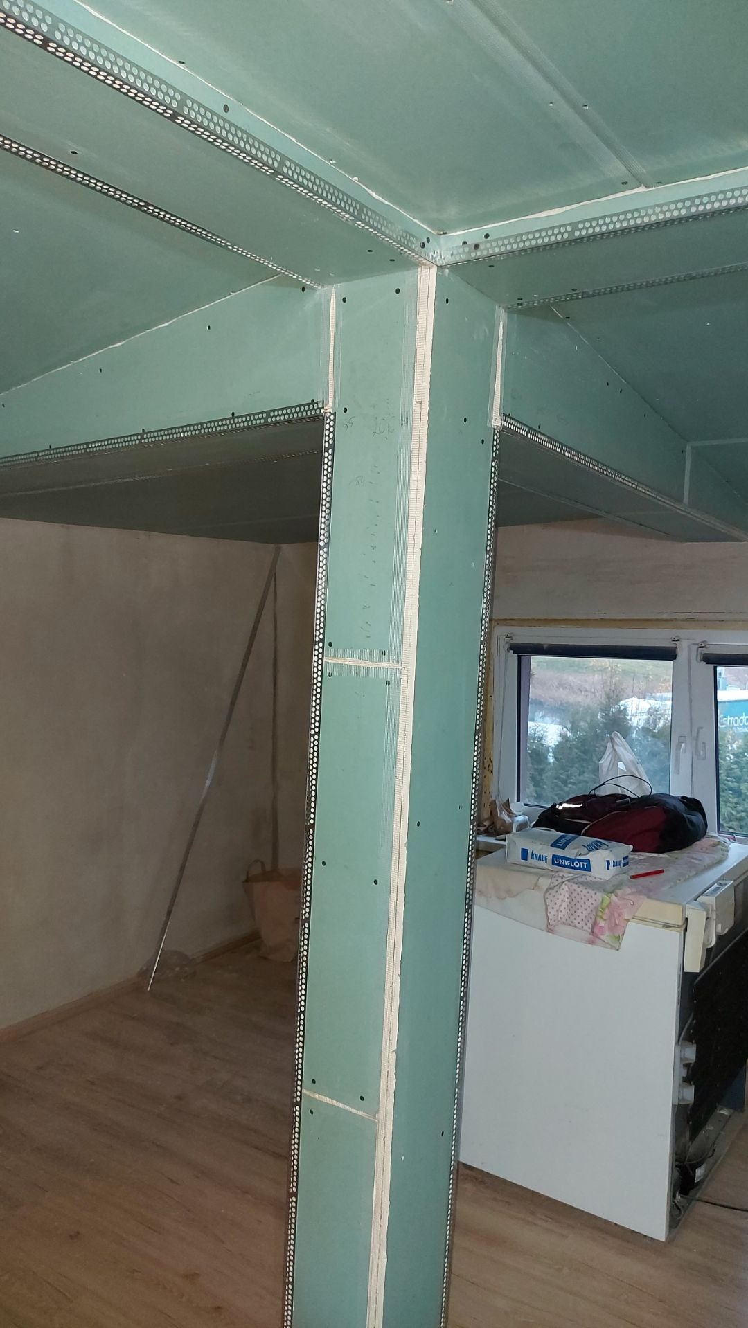 Konstrukcja z płyt gipsowo-kartonowych z profilami narożnikowymi, widoczna w surowym wnętrzu z drewnianą podłogą i elementami wyposażenia.