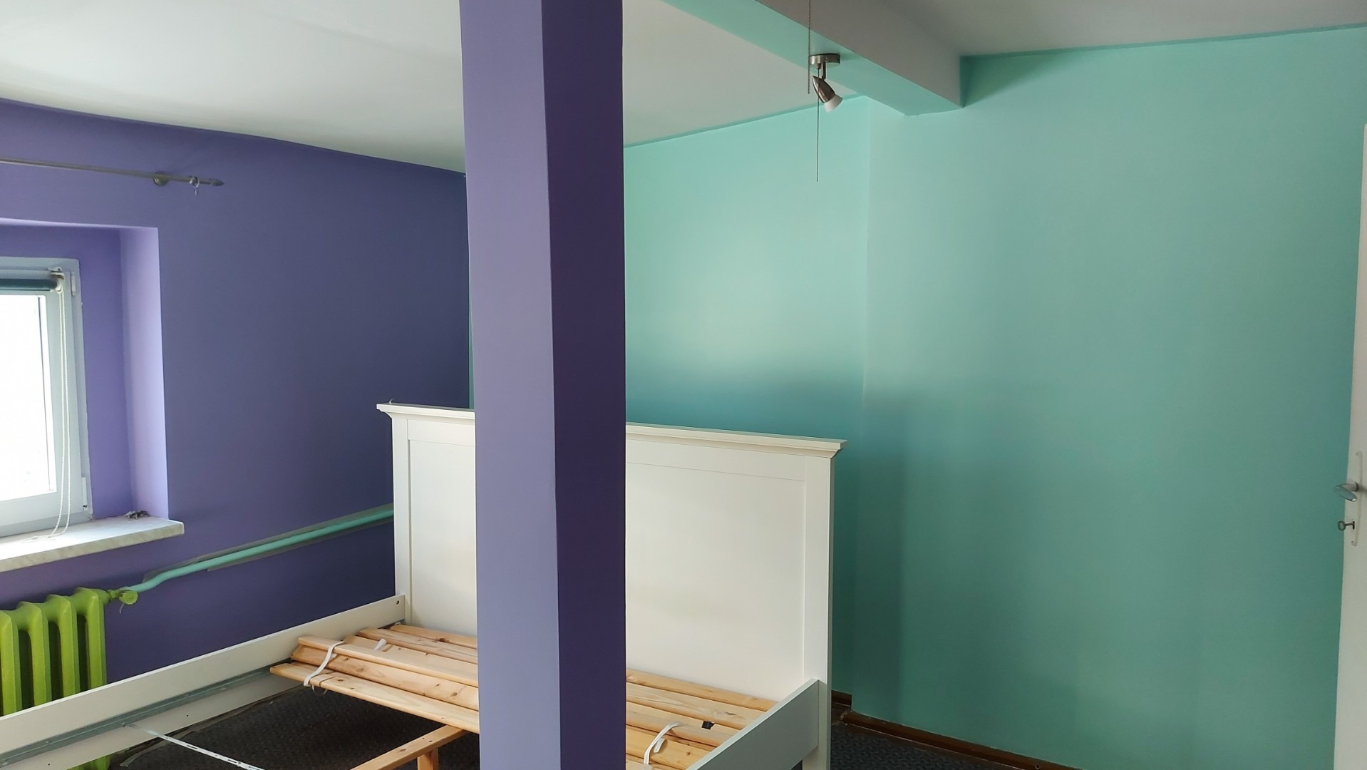 Pomalowane na fioletowo i miętowo ściany pokoju z częściowo zmontowanym białym łóżkiem, grzejnik w kolorze zielonym.