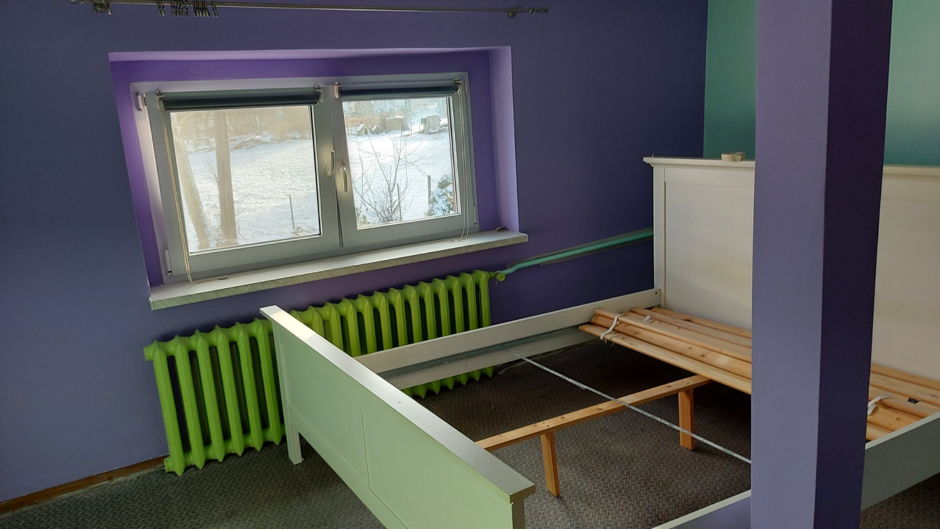 Fioletowy pokój z białym łóżkiem w trakcie montażu, widoczny zielony grzejnik pod oknem.