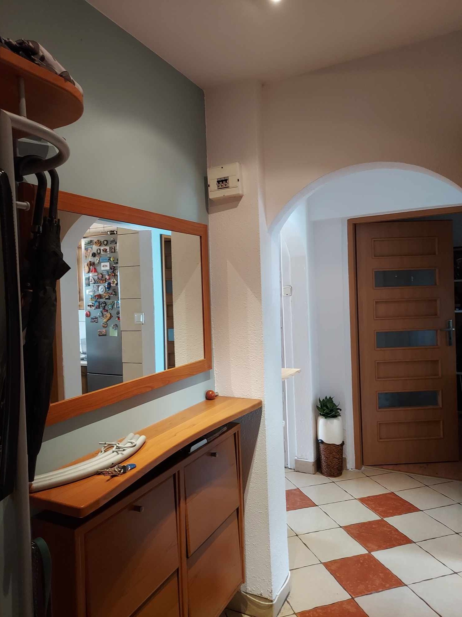 Przedpokój z szaro-niebieską ścianą, drewnianym lustrem i szafką na buty, podłoga w biało-czerwoną szachownicę, widoczny fragment kuchni w odbiciu lustra z magnesami na lodówce.