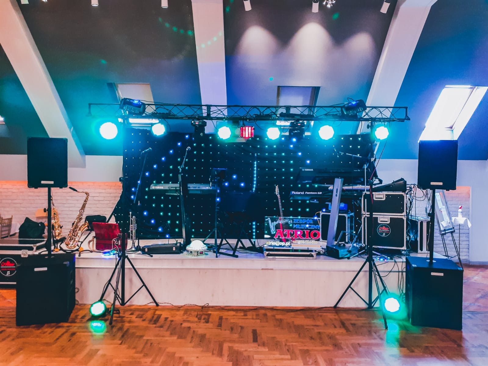 Scena z instrumentami muzycznymi, oświetleniem i nagłośnieniem, przygotowana do występu, widoczny saksofon, keyboardy Yamaha i Roland, oraz logo zespołu Atrio.