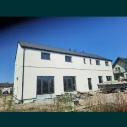 Nowoczesny, dwukondygnacyjny budynek z jasną elewacją i ciemnym dachem, duże okna balkonowe, widoczne materiały budowlane na zewnątrz, w tle błękitne niebo.