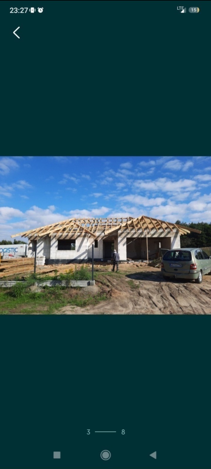 Niedokończony dom jednorodzinny z białego pustaka, widoczna konstrukcja dachu z drewnianych belek, teren wokół domu częściowo zagospodarowany, na podjeździe stoi zaparkowany samochód.