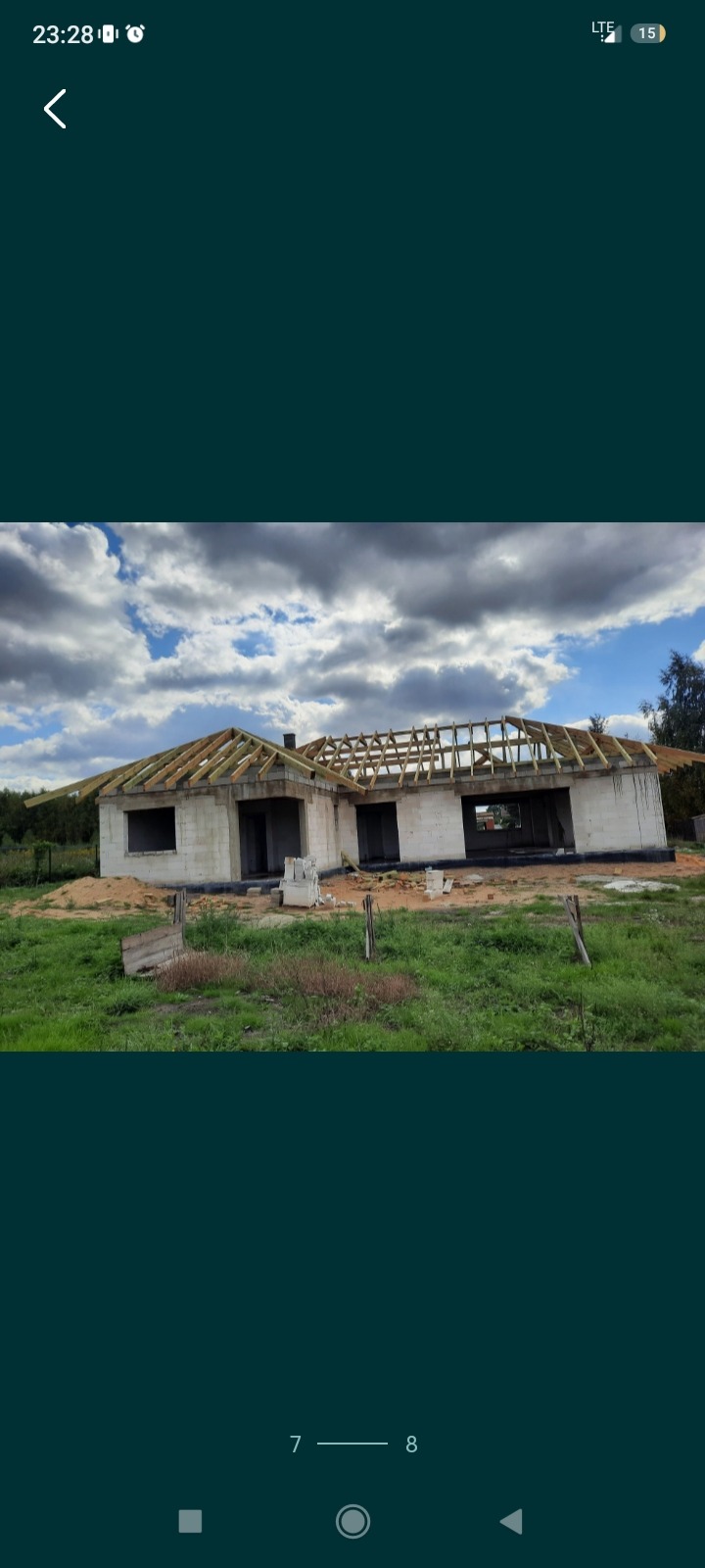 Niedokończony dom z bloczków betonowych w trakcie budowy dachu z drewnianą konstrukcją, widok z zewnątrz na tle zachmurzonego nieba i trawy.