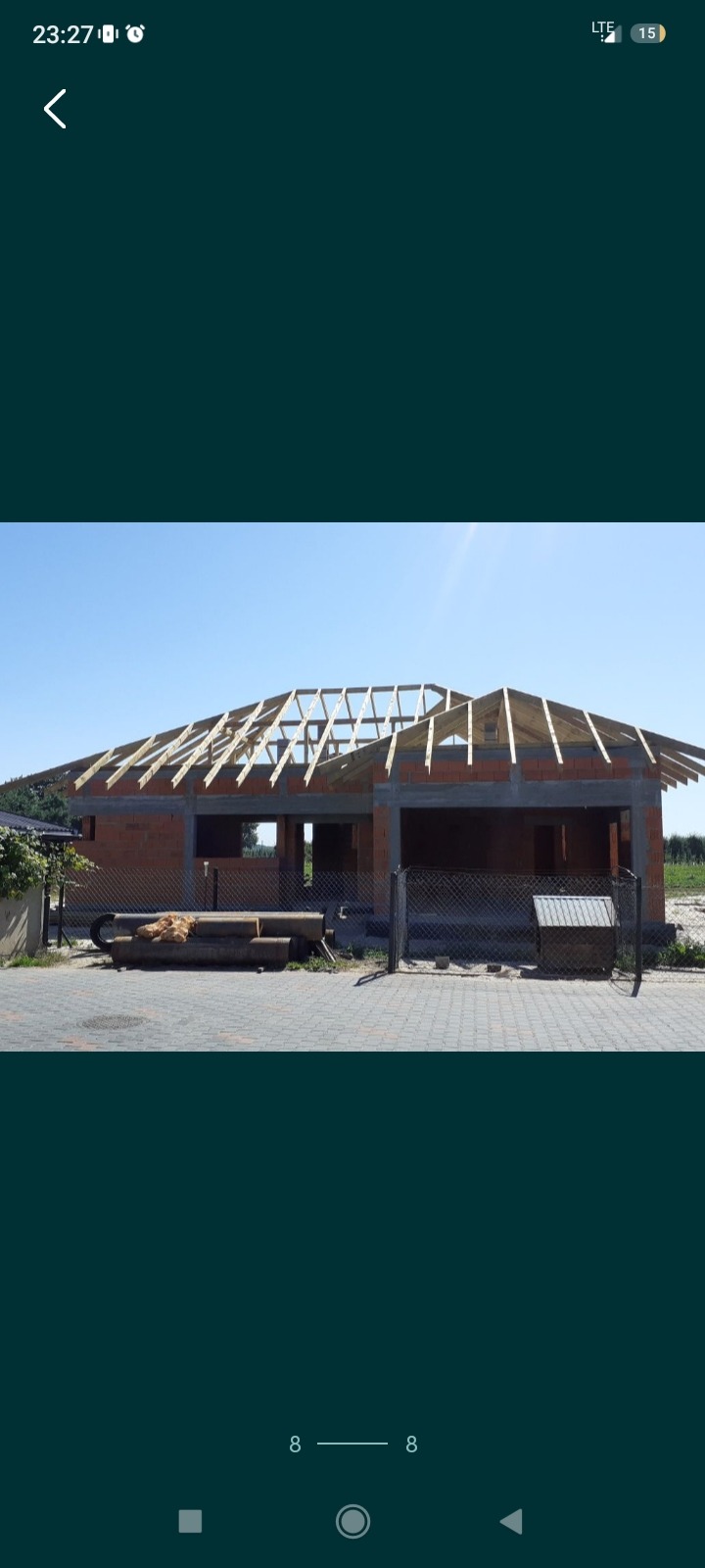 Budowa domu jednorodzinnego z czerwonej cegły, widoczny szkielet dachu z drewnianych belek, teren budowy zabezpieczony siatką, fragment zieleni w tle.