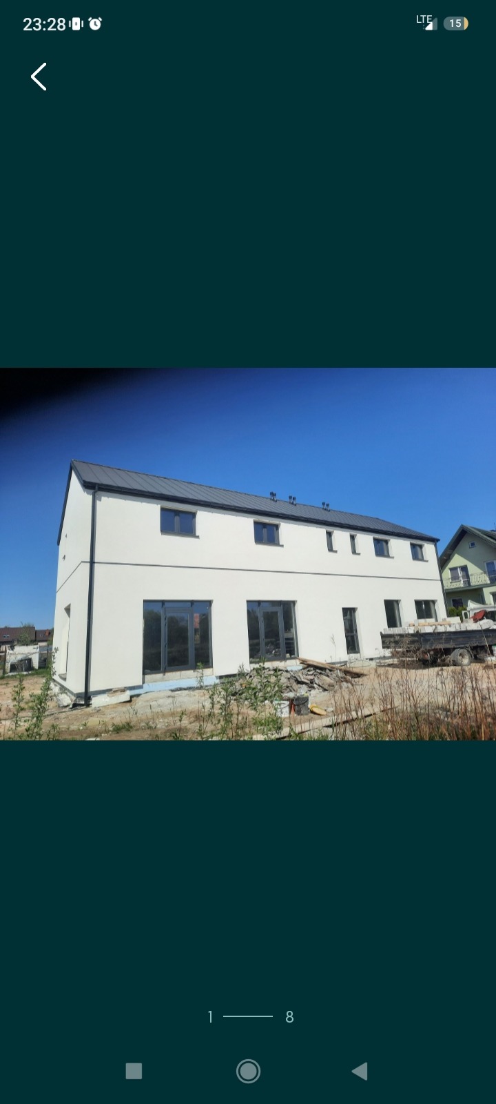 Nowoczesny, dwukondygnacyjny budynek z jasną elewacją i ciemnym dachem, duże okna balkonowe, widoczne materiały budowlane na zewnątrz, w tle błękitne niebo.