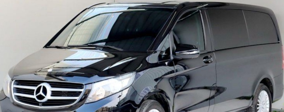 Błyszczący, czarny Mercedes-Benz Vito z przyciemnianymi szybami, zaparkowany w jasnym pomieszczeniu, widok z przodu i boku.