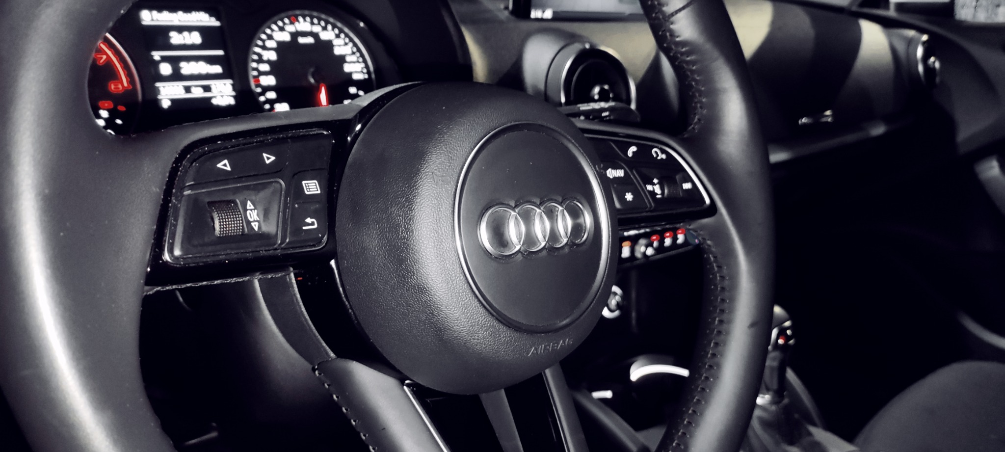 Wnętrze samochodu Audi z widoczną kierownicą, deską rozdzielczą i zegarami, ukazujące detale wykończenia i elementy sterowania.