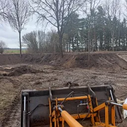 Żółta ładowarka na błotnistym placu budowy z widocznym wykopem i lasem w tle, pochmurne niebo.