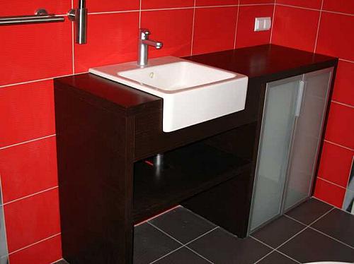 Nowoczesna łazienka z ciemnobrązową szafką pod umywalkę z półkami i przeszklonymi drzwiczkami, biała umywalka nablatowa, czerwone płytki na ścianie, czarne płytki na podłodze.