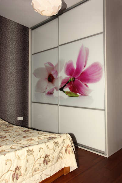 Biała szafa przesuwna z grafiką kwiatów magnolii, wkomponowana w aranżację sypialni z wzorzystą tapetą i beżową narzutą na łóżku.