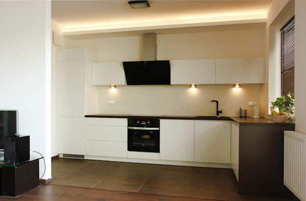 Nowoczesna, minimalistyczna kuchnia z białymi frontami i ciemnym blatem, oświetlona listwą LED pod sufitem i punktowymi lampkami podszafkowymi, widok z salonu.