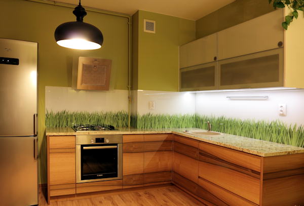 Kuchenna zabudowa z drewnianymi frontami, blatem w odcieniach beżu i zielonym akcentem w postaci naklejki z motywem trawy na ścianie nad blatem roboczym. Nad płytą grzewczą wisi drewniany okap,...