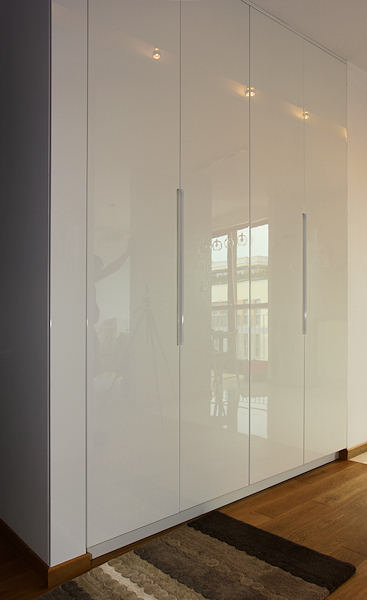 Biała, połyskująca szafa wnękowa z podłużnymi, metalowymi uchwytami na tle drewnianej podłogi i dywanika w odcieniach brązu, z widocznym odbiciem wnętrza pokoju.