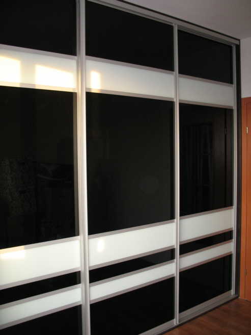 Trzydrzwiowa szafa wnękowa z przesuwnymi drzwiami Raumplus, z czarnymi panelami i białymi poziomymi pasami, widoczna na tle drewnianej podłogi i fragmentu brązowych drzwi.