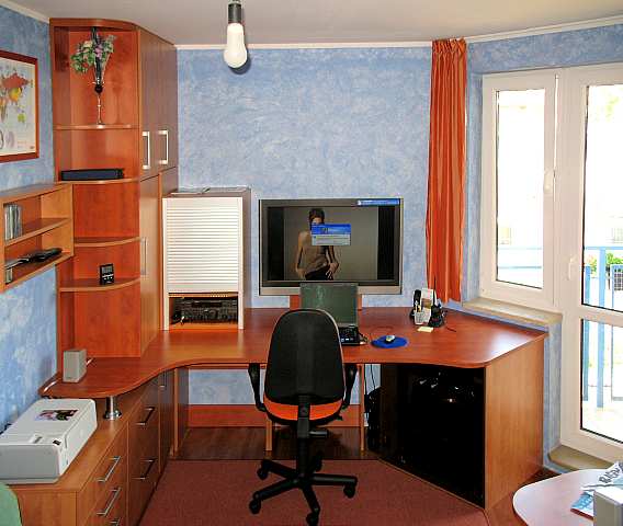 Ergonomiczne biuro domowe z narożnym biurkiem z drewna, szafkami, półkami, roletą, komputerem i pomarańczowym akcentem na krześle biurowym, doświetlone wiszącą żarówką.