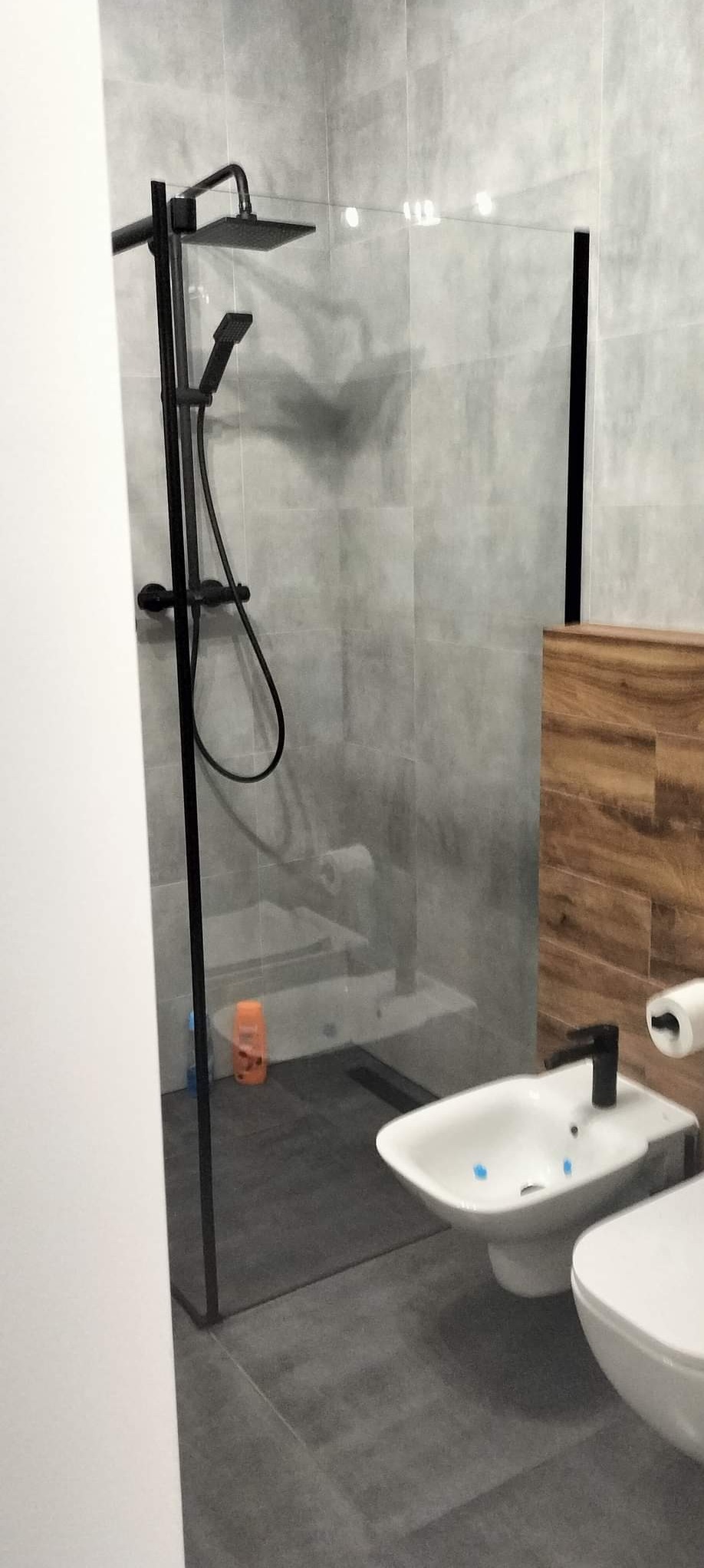 Nowoczesna łazienka z szarą glazurą, czarną armaturą, kabiną prysznicową typu walk-in, bidetem i toaletą.