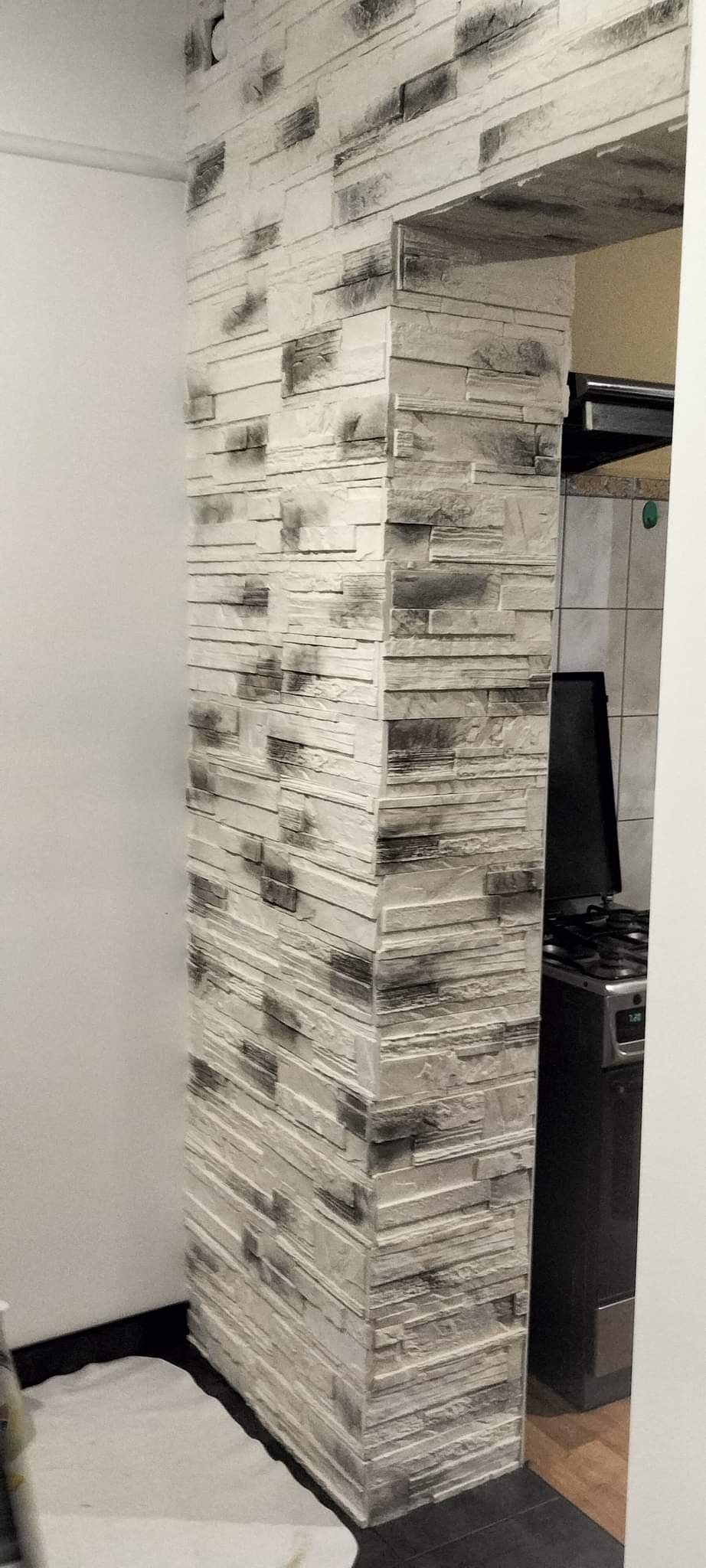 Wykończenie ściany imitujące kamień w odcieniach szarości i bieli, tworzące dekoracyjny element architektoniczny przy wejściu do kuchni z widoczną kuchenką gazową.