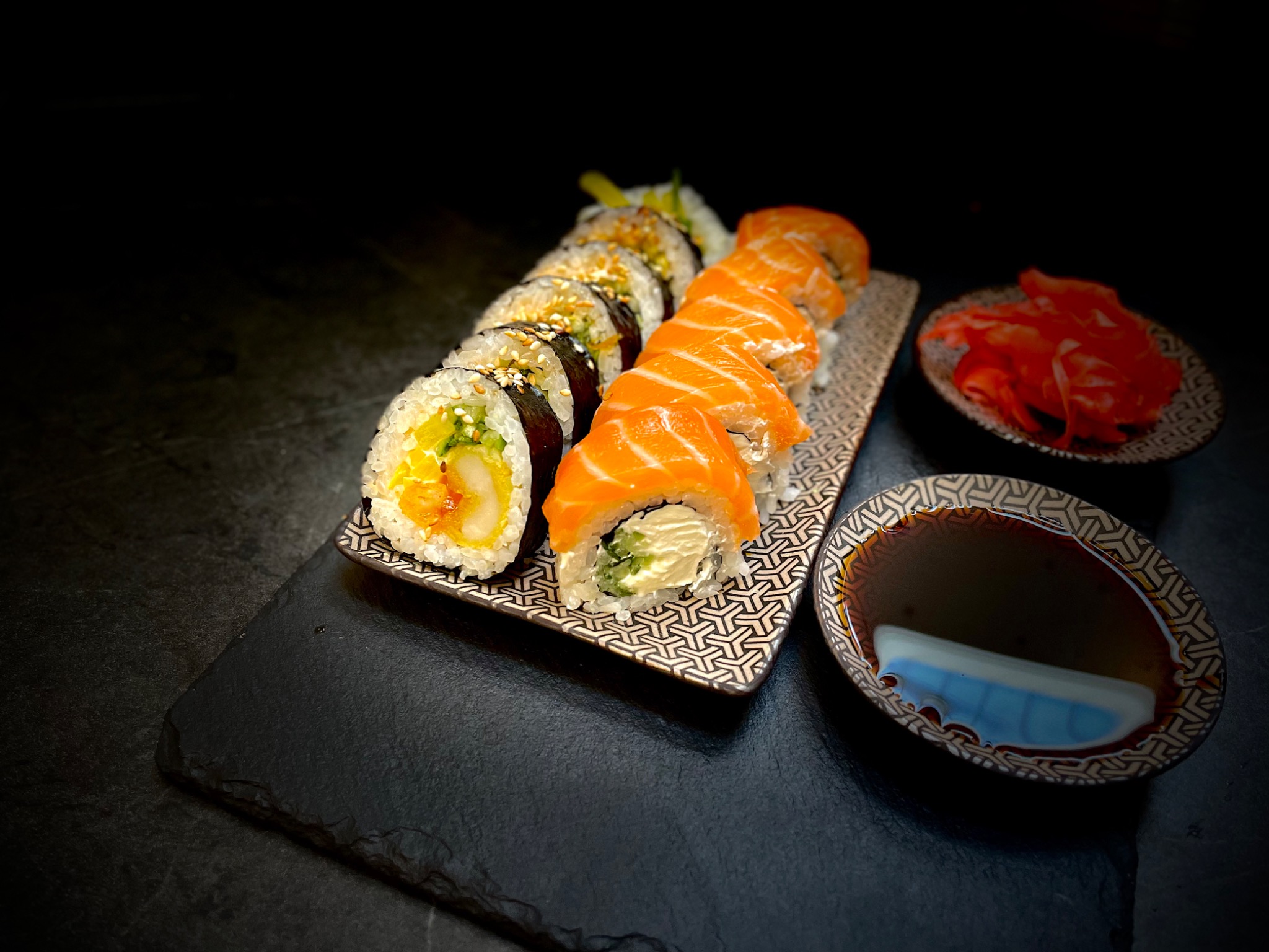 Zestaw sushi z łososiem, krewetką i warzywami, podany na ozdobnym talerzu z sosem sojowym i marynowanym imbirem na czarnym kamiennym tle.