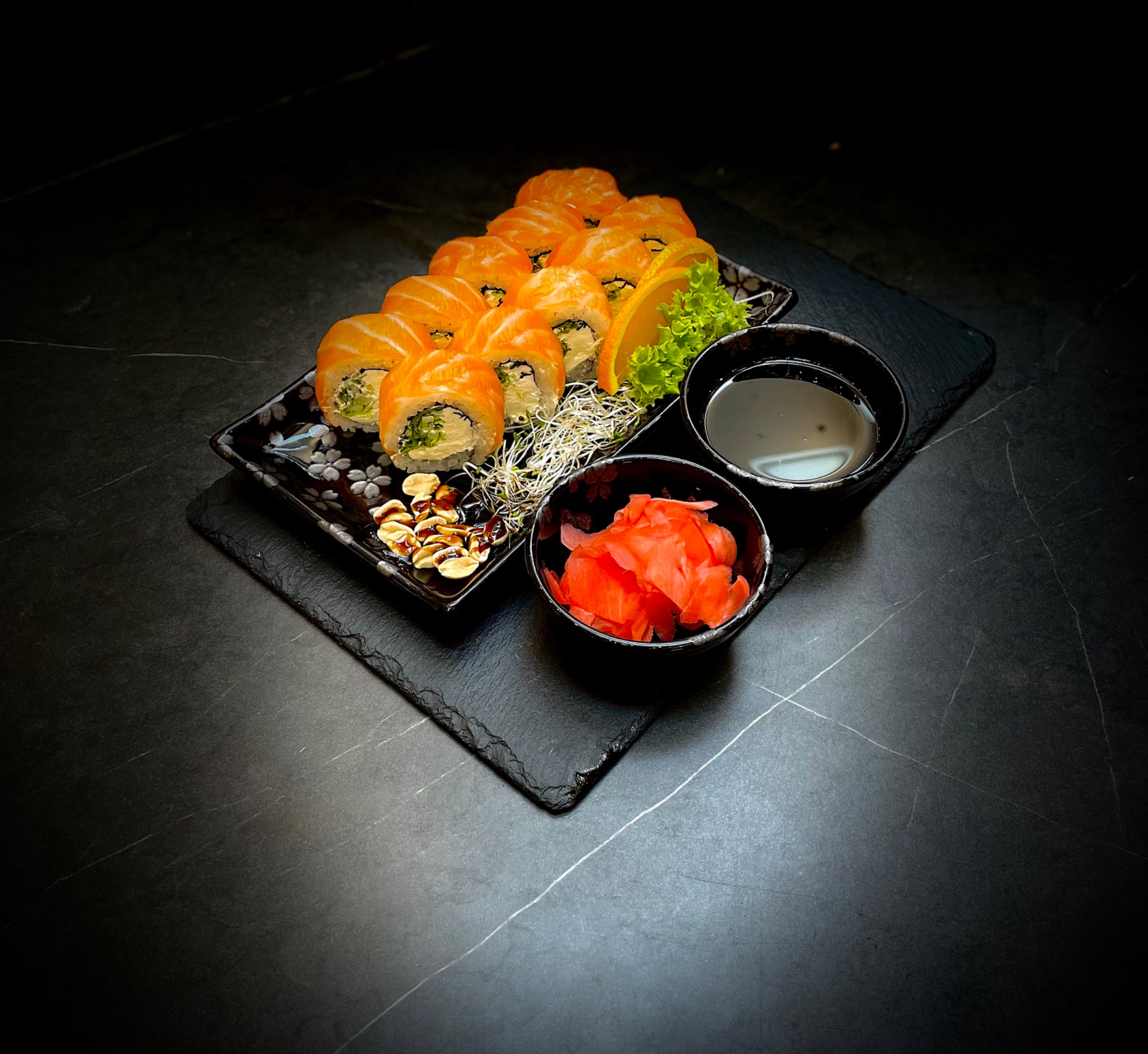 Ułożone na czarnym, kwadratowym talerzu sushi z łososiem, z zielonym nadzieniem, dekorowane kiełkami, plasterkiem pomarańczy i sałatą, obok miseczki z sosem sojowym i imbirem na czarnej, kamiennej...
