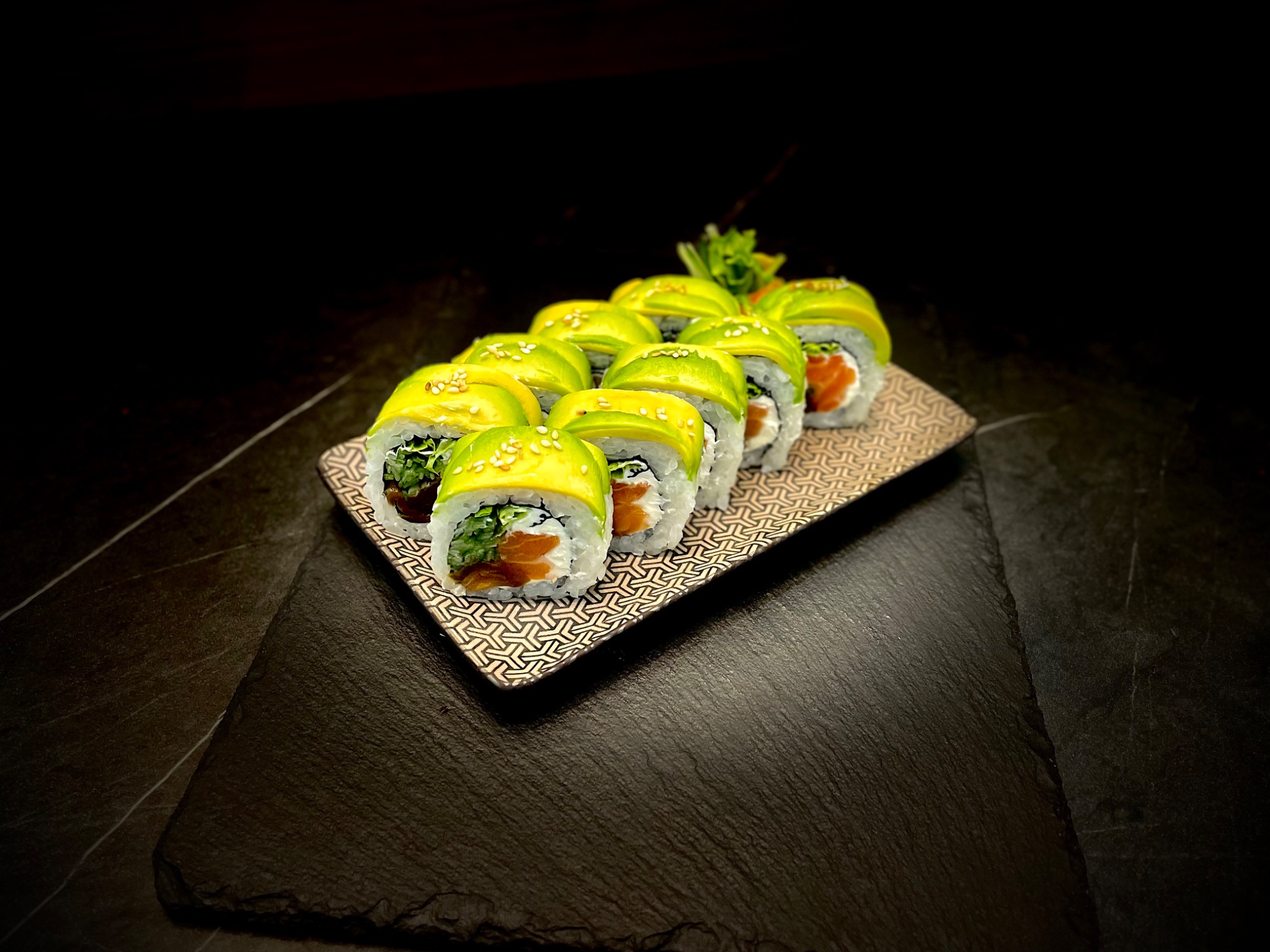 Rząd rolek sushi z łososiem, awokado i sezamem, ułożonych na wzorzystej tacy na ciemnym, kamiennym tle.