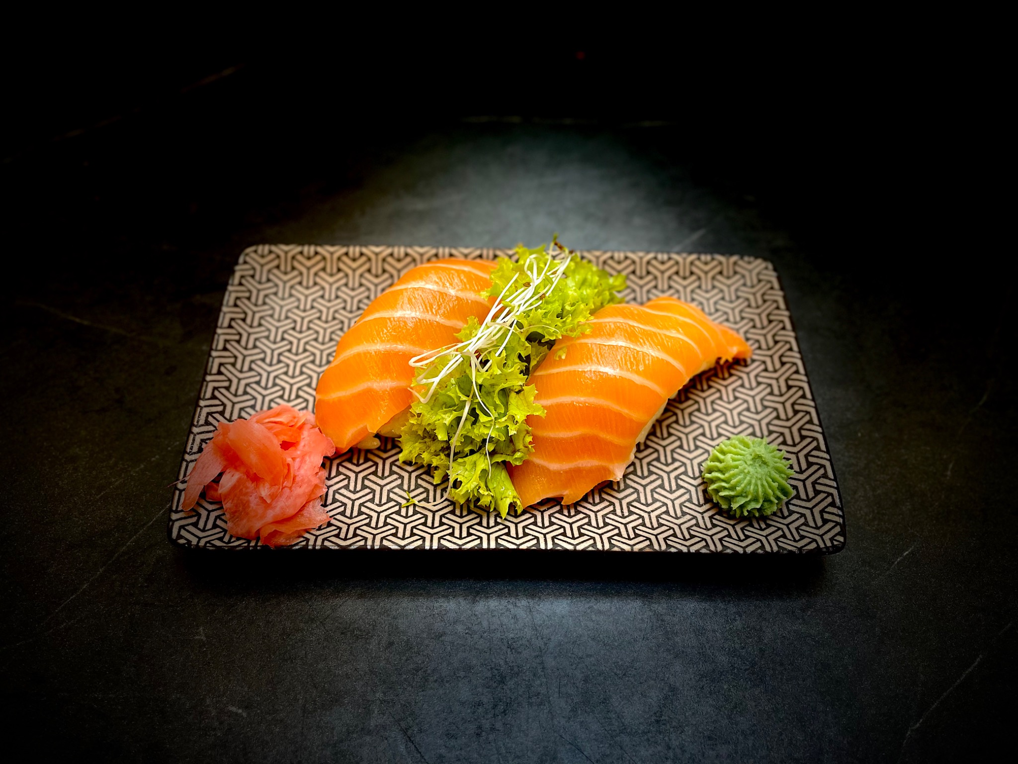 Dwa kawałki nigiri z łososiem, dekorowane sałatą i kiełkami, podane na wzorzystym talerzu z imbirem i wasabi na ciemnym tle.