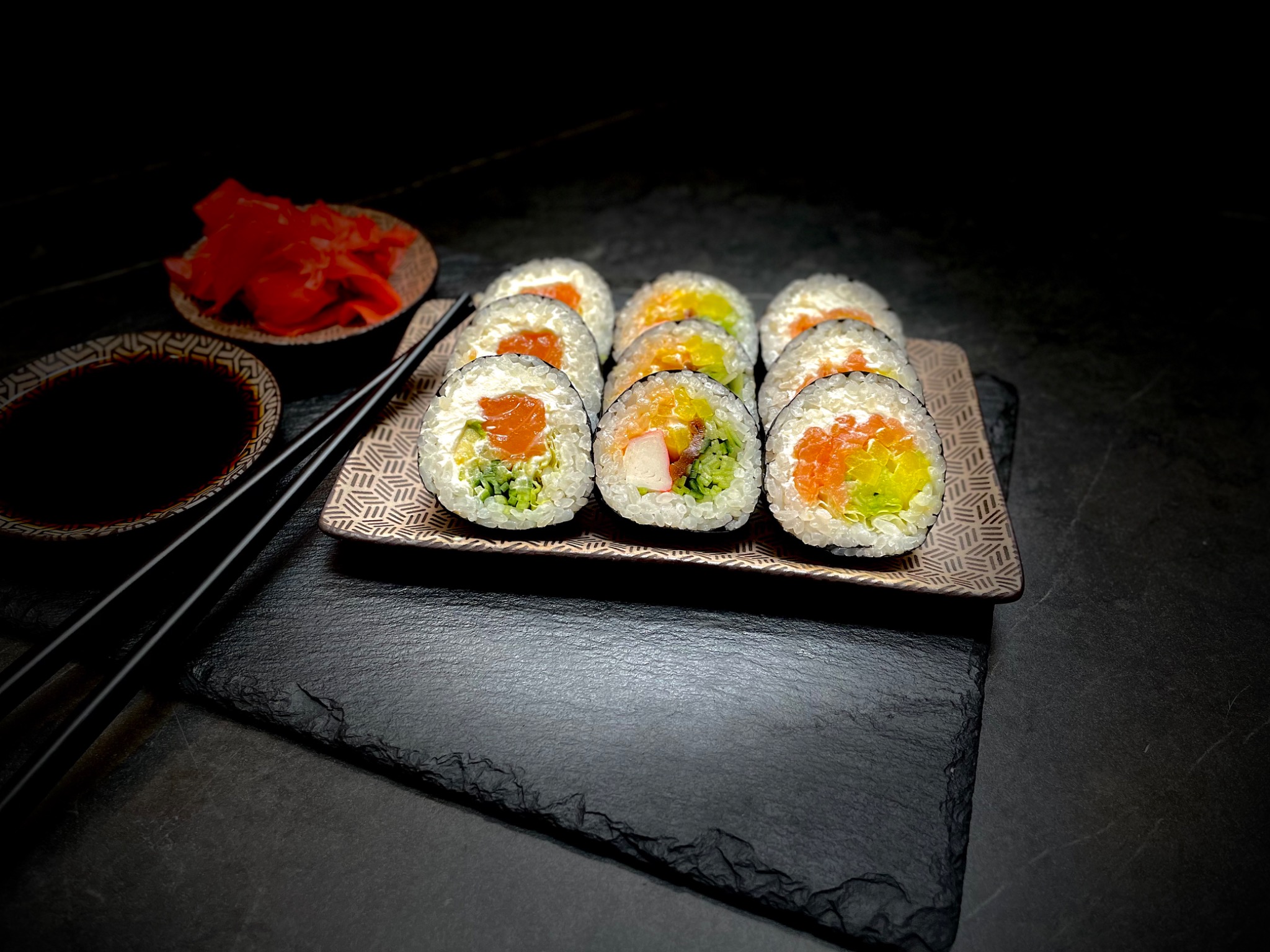 Ułożone na wzorzystym talerzu rolki sushi z łososiem, warzywami i białym serem, podane z imbirem i sosem sojowym, na czarnym kamiennym tle.