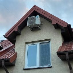 M.M KLIMA Mateusz Miechówka - Zewnętrzna jednostka klimatyzacji zamontowana na ścianie szczytowej domu, widoczna pod okapem dachu, z oknem poniżej. Na ścianie widoczna faktura tynku.