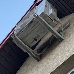 M.M KLIMA Mateusz Miechówka - Zewnętrzna jednostka klimatyzacji zamontowana na wsporniku na elewacji budynku pod okapem dachu.