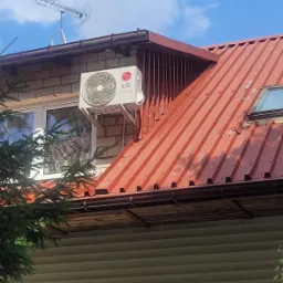 Zewnętrzna jednostka klimatyzacji LG przymocowana do ściany budynku z cegły, widoczna antena telewizyjna na dachu.