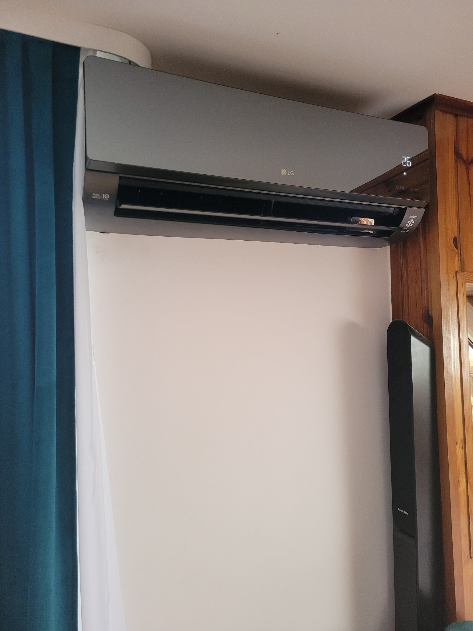 Szara klimatyzacja LG zamontowana na ścianie w pokoju z zasłonami i drewnianymi elementami wykończenia, wyświetlająca temperaturę 26 stopni Celsjusza.